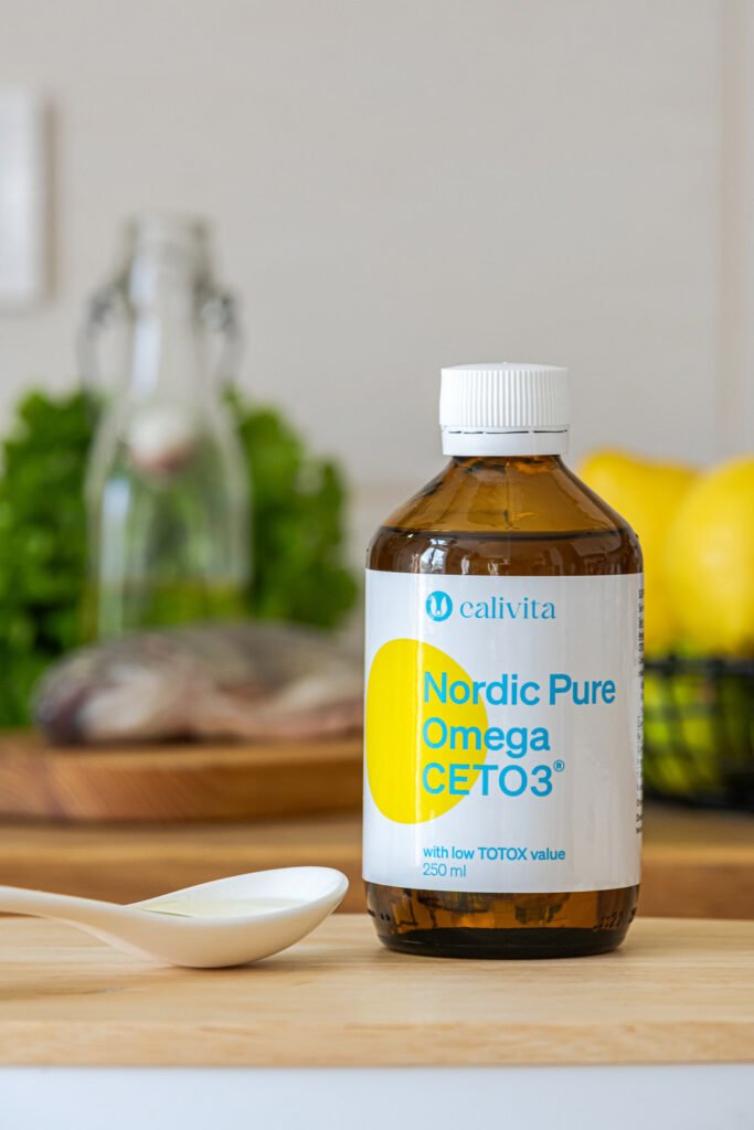 Nordic Pure Omega CETO3 CaliVita z łyżeczką, w tle świeży śledź, zioła i cytryny