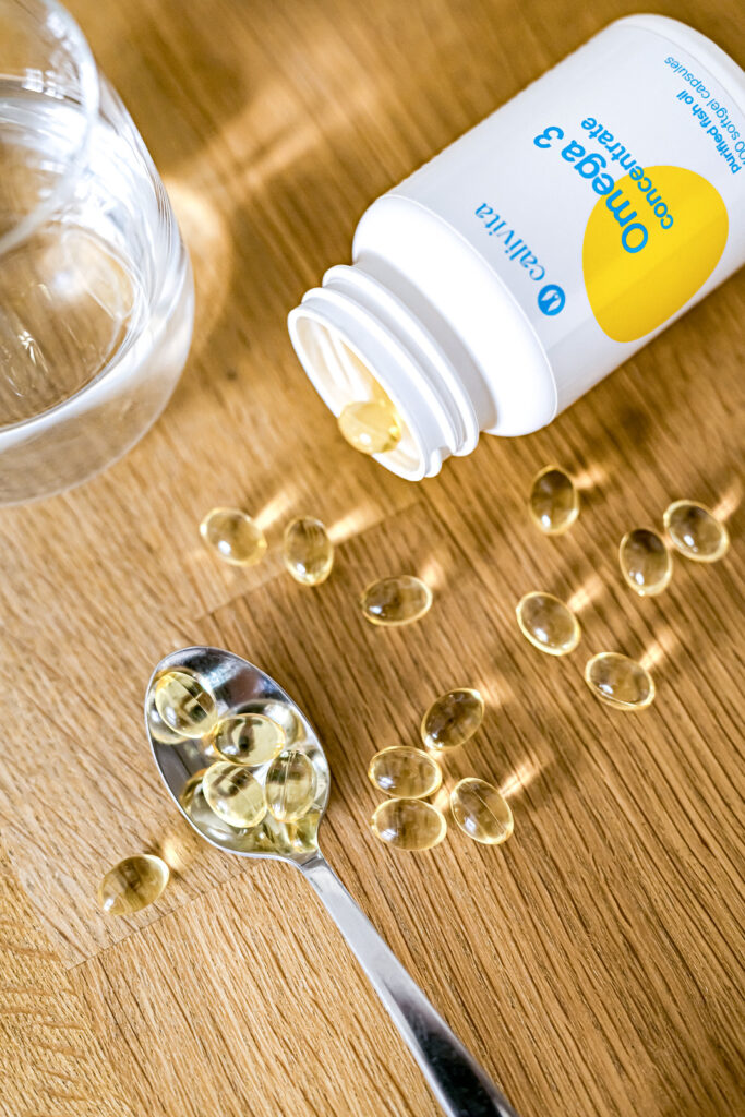 Małe kapsułki żelowe Omega 3 Concentrate CaliVita na łyżce, obok szklanka wody